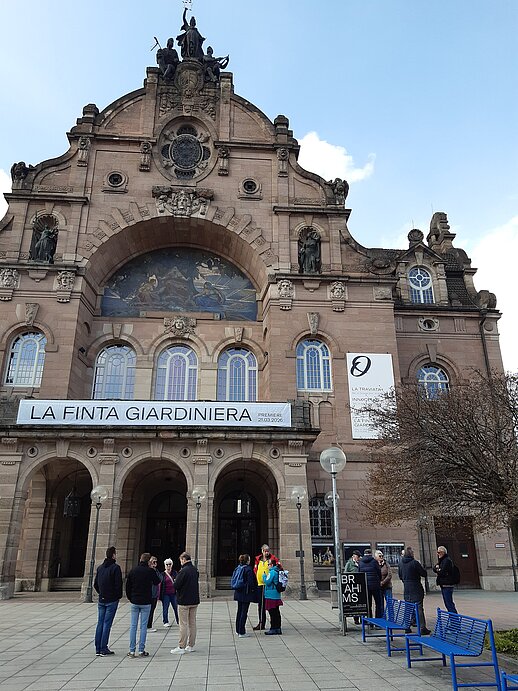 Außenansicht des großen, historischen Staatstheatergebäudes in Nürnberg aus hellem Stein. Die Fassade ist reich verziert mit Figuren, Bögen und einem großen Rundfenster. Über dem Eingang hängt ein langes Banner mit dem Text „La Finta Giardiniera“. Vor dem Gebäude stehen mehrere kleine Gruppen von Menschen auf einem Platz. Rechts sind eine Bank, ein Baum und eine Laterne zu sehen, darüber ein blauer Himmel.