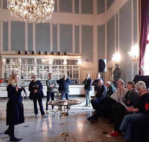 Großer, heller Saal mit hohen Wänden und Kronleuchter. Im Hintergrund eine Bar mit Regalen voller Gläser. Links steht eine Person und spricht zu einer Gruppe. Rechts sitzen mehrere Menschen nebeneinander und hören zu, einige stehen dahinter. In der Mitte stehen kleine runde Tische.